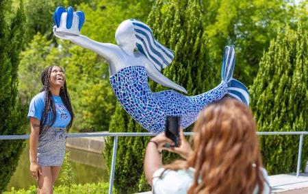 a student wearing an blue shirt is standing under the odu norfolk mermaid while another student takes her photo